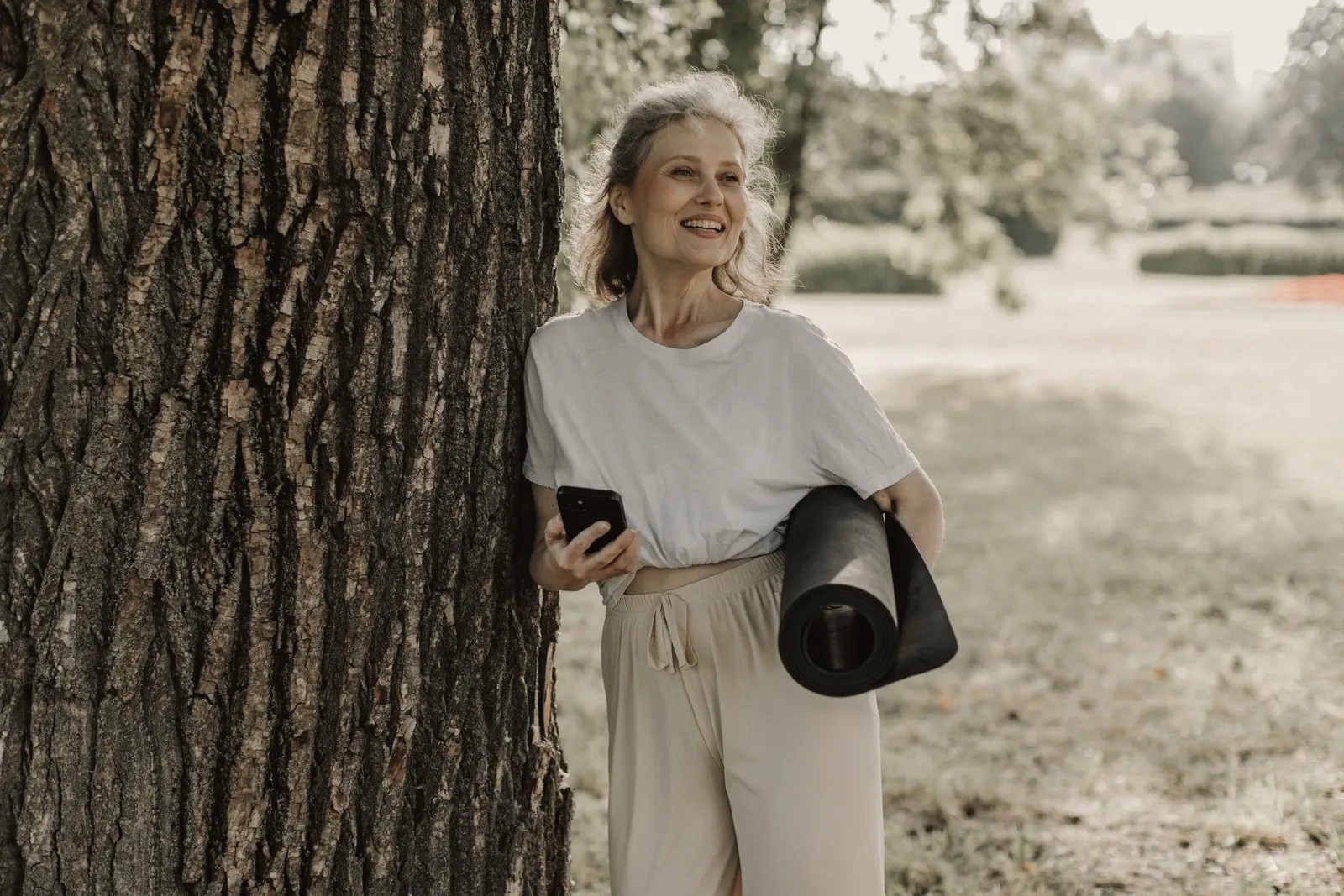 Mujer madura sonriente apoyada en un árbol, con una esterilla de ejercicio bajo el brazo