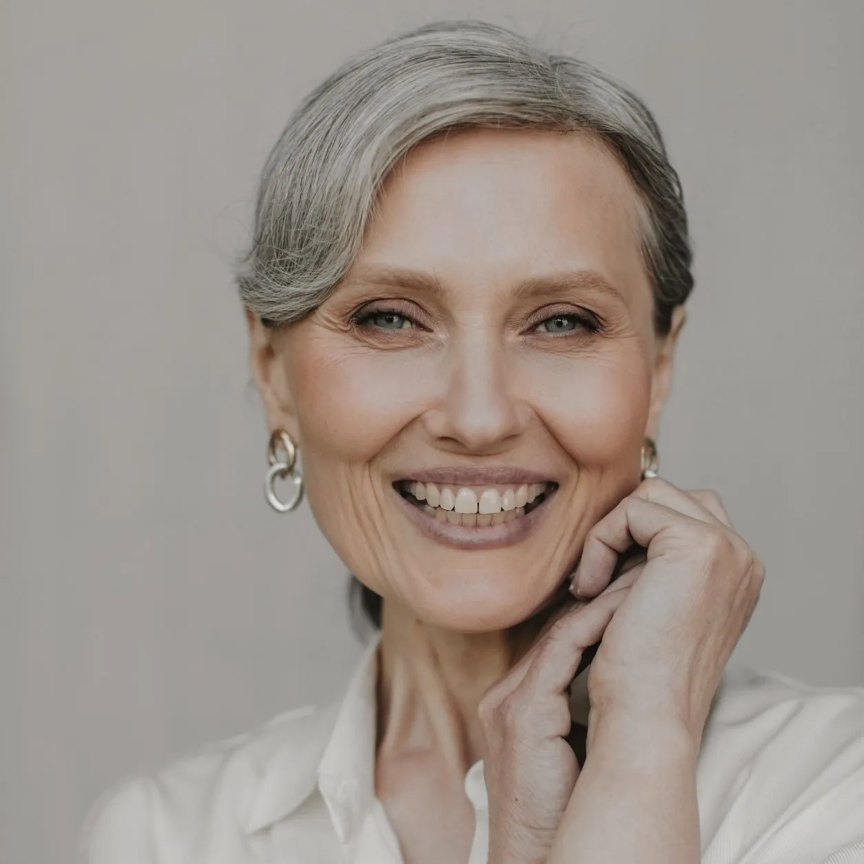Mujer de pelo gris recogido sonriendo, con las manos junto a la cara y la piel luminosa